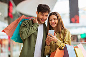 Couple Shopping Using Phone Application Holding Shopper Bags In Mall
