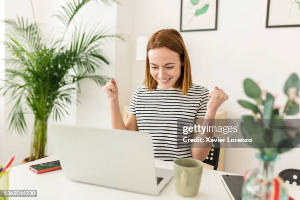 cheerful young woman celebrating her achievement while reading good news on laptop from home office - business woman cheering stockfoto's en -beelden