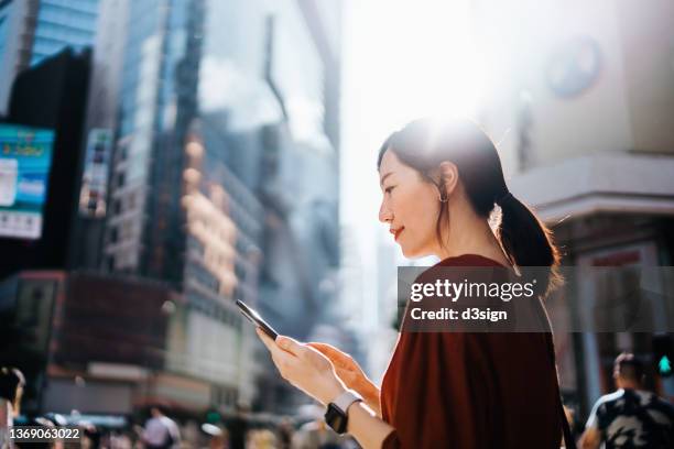 side profile of confident young asian businesswoman using smartphone on the go, walking in busy downtown district against crowd of pedestrians and urban skyscrapers with morning sunlight. female leadership, girl power, business on the go concept - one person in crowd stock pictures, royalty-free photos & images