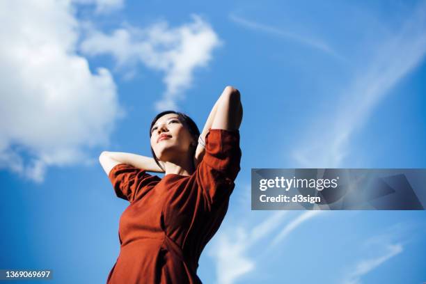 low angle portrait of young asian woman stretching arms behind her head, setting herself free and feeling relived. standing against beautiful clear blue sky enjoying sunlight. freedom in nature. connection with nature - low confidence stock pictures, royalty-free photos & images