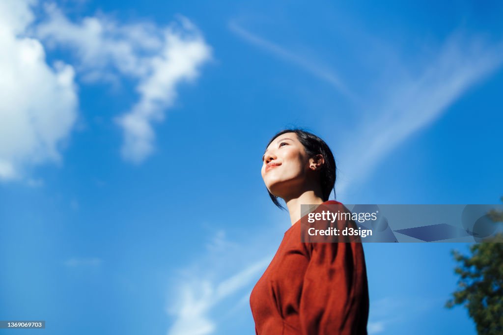 Low angle portrait of beautiful smiling young Asian woman standing against beautiful clear blue sky, looking up to sky enjoying nature and sunlight. Freedom in nature. Connection with nature