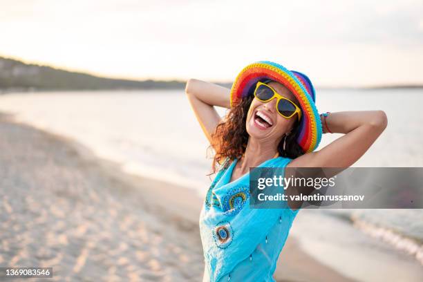 portrait of a smiling woman with colorful hat, enjoying the day on the beach, posing during golden hour - golden hour beach stock pictures, royalty-free photos & images