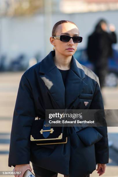 Guest wears black sunglasses, gold earrings, a black t-shirt, a black oversized Re-Nylon puffer jacket from Prada, a black suede with gold borders...