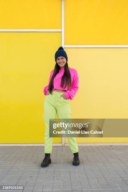 18-year-old woman wearing a pink sweater, blue cap and green jeans on a yellow wall. - pink pants stock-fotos und bilder