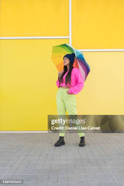 18-year-old woman wearing a pink sweater, blue cap, green jeans and a rainbow-colored umbrella on a yellow wall. - fondo amarillo stock pictures, royalty-free photos & images