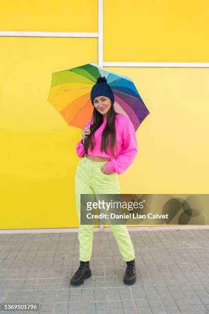 18-year-old woman wearing a pink sweater, blue cap, green jeans and a rainbow-colored umbrella on a yellow wall. - fondo amarillo stock pictures, royalty-free photos & images