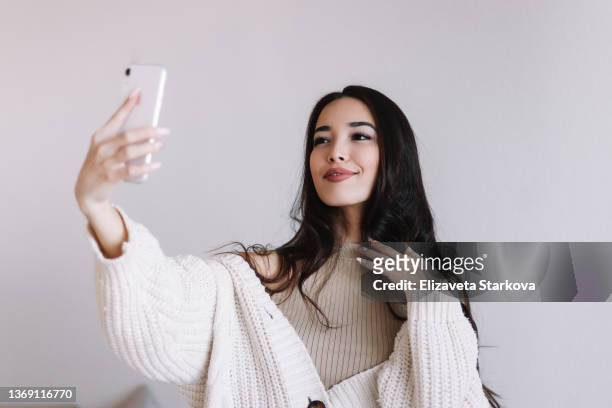 a young asian millennial woman takes a selfie and communicates on the phone using wireless technology. large portrait of a cheerful smiling girl chatting on an online video on a white isolated background - selfi stock-fotos und bilder