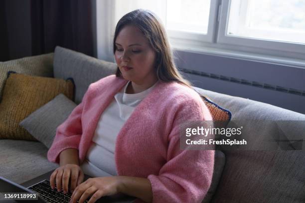 beautiful overweight woman sitting on the sofa in her living room and browsing the web on a laptop - beautiful fat women stock pictures, royalty-free photos & images