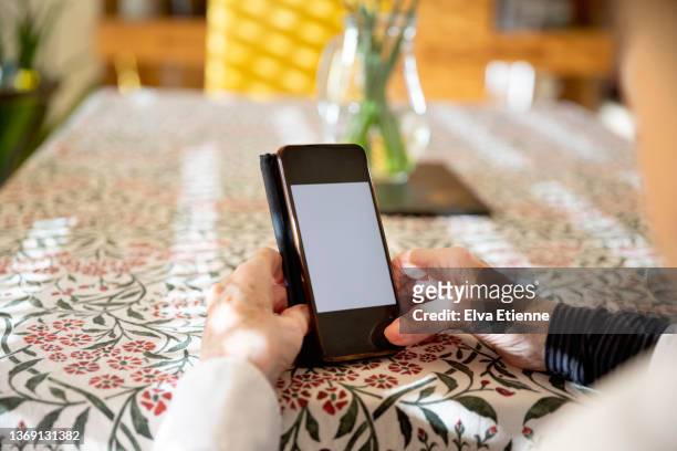 over the shoulder view of senior woman using mobile phone with a blank white screen, whilst sitting at a dining table - hand back lit stock pictures, royalty-free photos & images