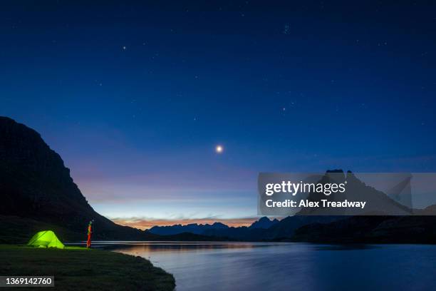 a man standing beside his tent looks up at a star filled night sky above a lake - pyrenees stock-fotos und bilder