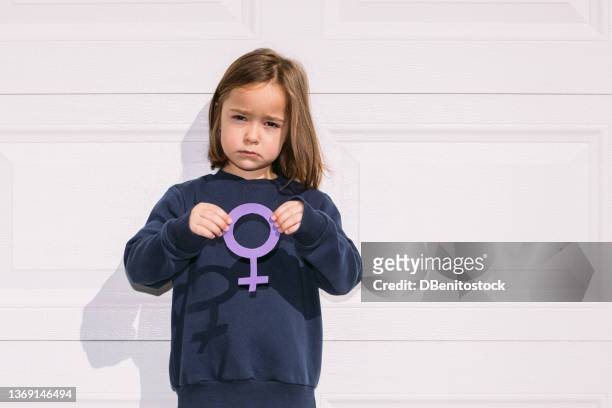 little blonde-haired girl holding purple female symbol on white garage door. concept of women's day, empowerment, equality, inequality, activism and protest. - violet manners stock pictures, royalty-free photos & images