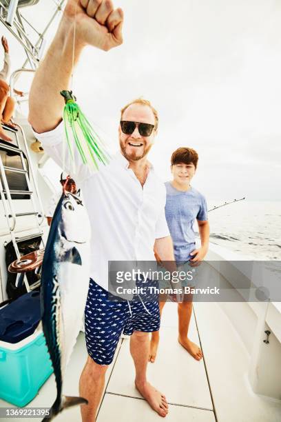 wide shot of smiling father holding up fish on stern of sport fishing boat while on trip with son - fang stock-fotos und bilder
