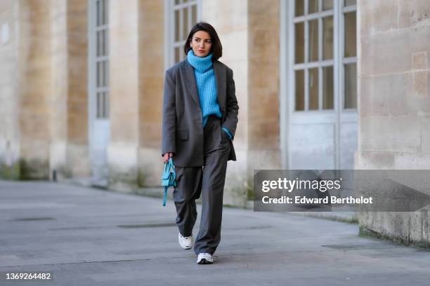 Alexandra Pereira wears earrings, a blue ribbed wool oversized turtleneck pullover, a gray oversized blazer jacket, matching gray oversized suit...