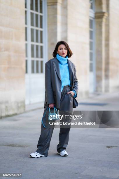 Alexandra Pereira wears earrings, a blue ribbed wool oversized turtleneck pullover, a gray oversized blazer jacket, matching gray oversized suit...