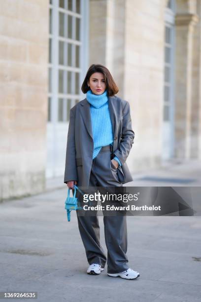 Alexandra Pereira wears earrings, a blue ribbed wool oversized turtleneck pullover, a gray oversized blazer jacket, matching gray oversized suit...
