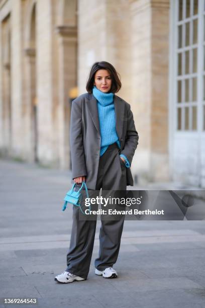 Alexandra Pereira wears earrings, a blue ribbed wool oversized turtleneck pullover, a gray oversized blazer jacket, matching gray oversized suit...