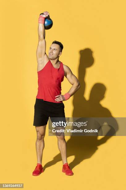 athletic man in red sportswear lifting kettle bell overhead against yellow background - handsome bodybuilders fotografías e imágenes de stock