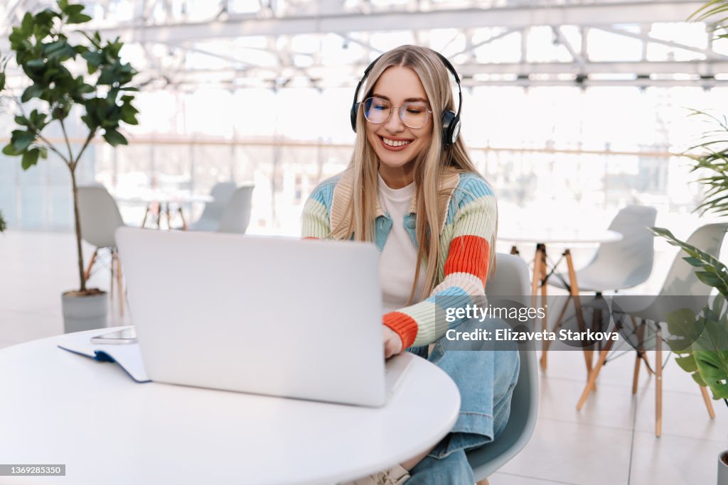 A young female gamer with headphones is playing using a laptop and listening to music while sitting at a table. A smiling girl student in glasses and casual clothes is studying online using wireless technology and listening to a scientific podcast