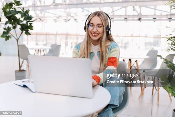 a young female gamer with headphones is playing using a laptop and listening to music while sitting at a table. a smiling girl student in glasses and casual clothes is studying online using wireless technology and listening to a scientific podcast - media profession for women stock pictures, royalty-free photos & images