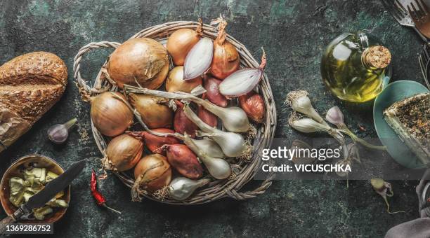 various onion in wooden basket at rustic dark kitchen table with baguette, garlic, olive oil and cheese - cebolla fotografías e imágenes de stock