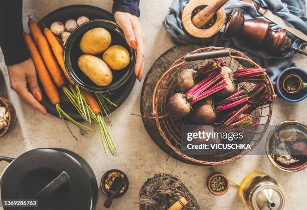 women hands holding bowl and plate with potatoes and carrots at white table with kitchen utensils, spices and beetroot - kartoffel wurzelgemüse stock-fotos und bilder