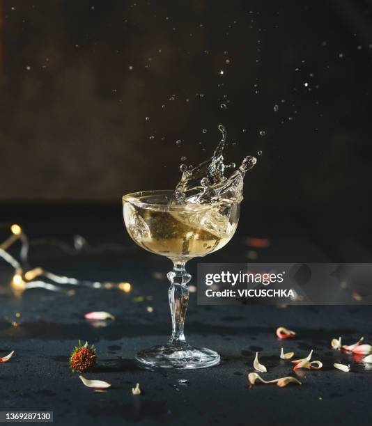 champagne glass with splashing beverage at dark kitchen table with flower petals and lightchain at wall background - champagne stock-fotos und bilder