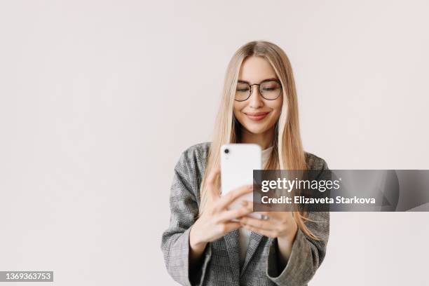 a young woman in glasses and casual clothes communicates online via video chat using a mobile phone on a white background. sociable student millennial blogger talking and looking at the phone. place for text - taking selfie white background stockfoto's en -beelden