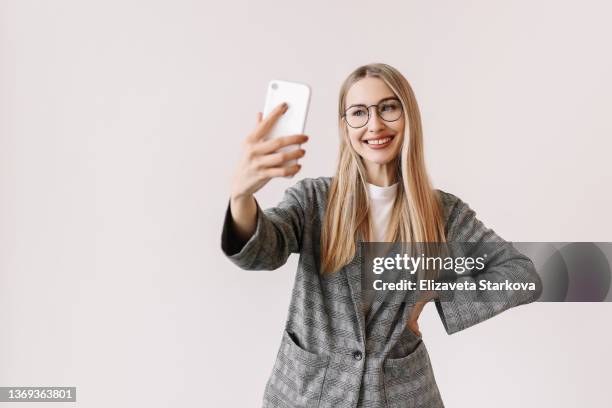 a young woman in glasses and casual clothes communicates online via video chat using a mobile phone on a white background. sociable student millennial blogger talking and looking at the phone. place for text - taking selfie white background stockfoto's en -beelden