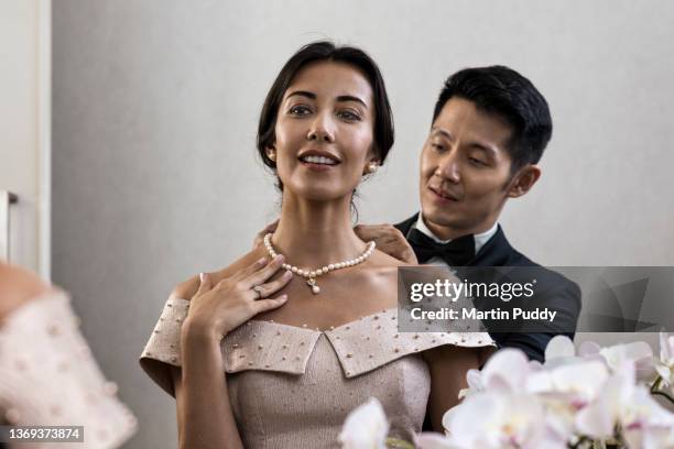 young multiracial couple at home wearing evening wear, adorning necklace in front of mirror - avondkledij stockfoto's en -beelden