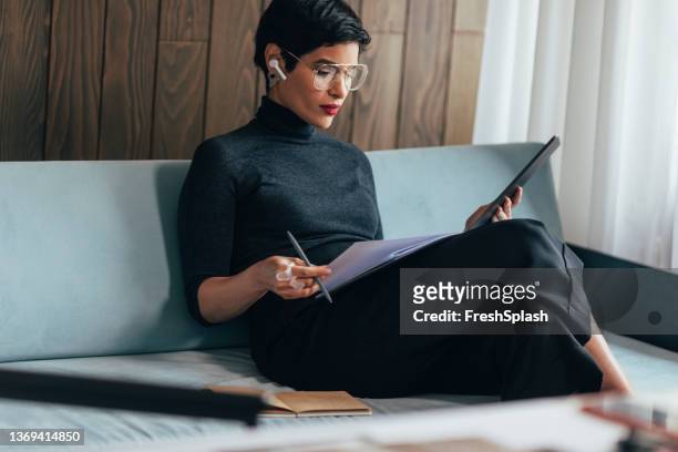 beautiful young businesswoman sitting on the couch in her office reading a report and using a digital tablet - business people and paper imagens e fotografias de stock