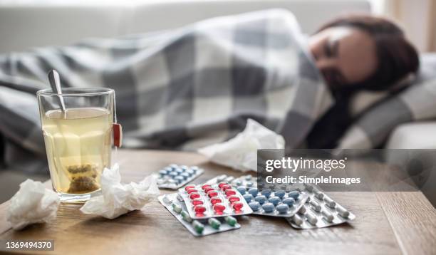 woman covered in blanket sleeps at home when feeling sick, having hot tea, used tissues and pills on the table next to her. - winter blues stock pictures, royalty-free photos & images