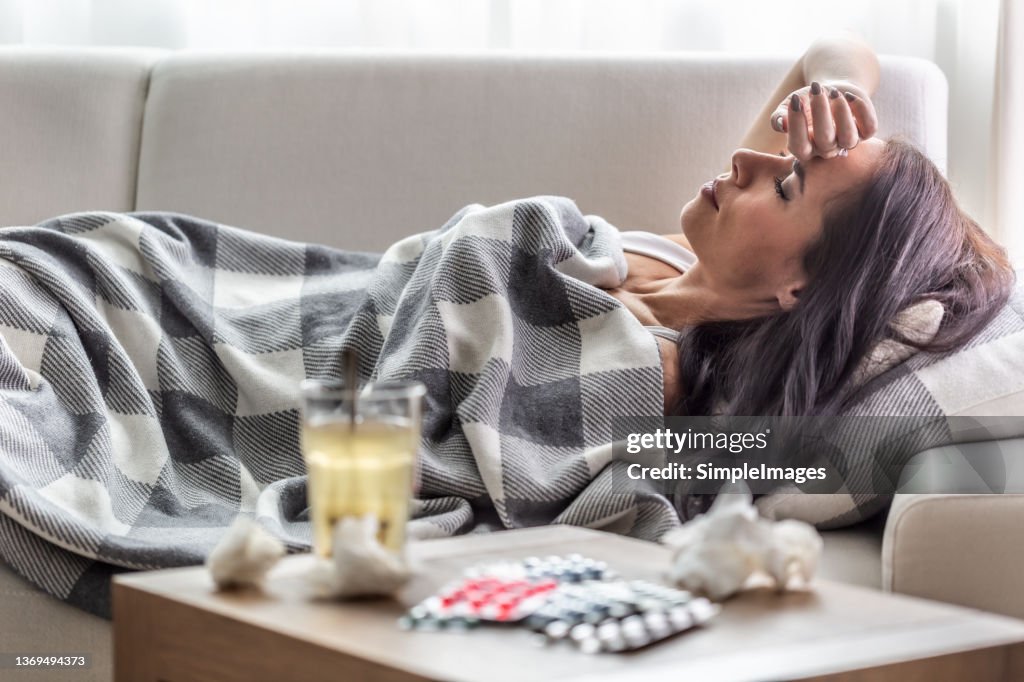 Woman having symptoms of Covid-19 lies covered in blanket in isolation with handerchiefs and pills next to her.