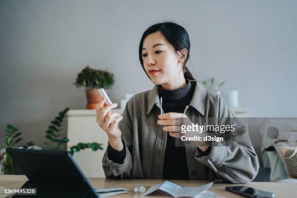 young asian woman holding a covid-19 rapid lateral flow test kit and a nasal swab, carrying out a coronavirus rapid self test at home. she is having a video call appointment on digital tablet with her family doctor for advice and consultation - video reviewed stock pictures, royalty-free photos & images