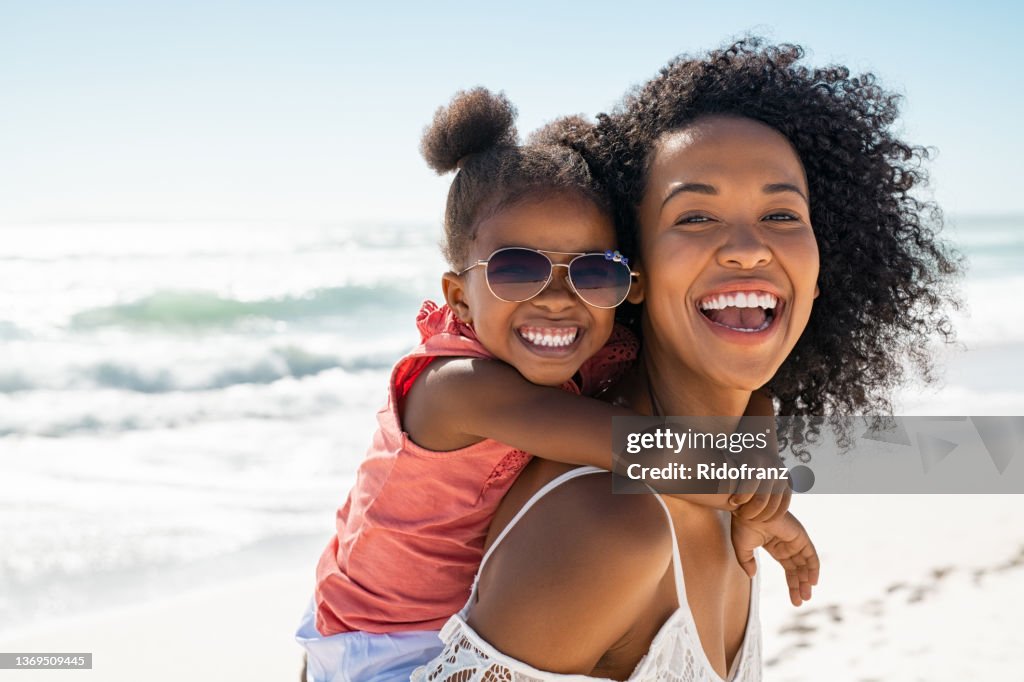 Happy young mother giving laughing daughter piggyback ride at beach