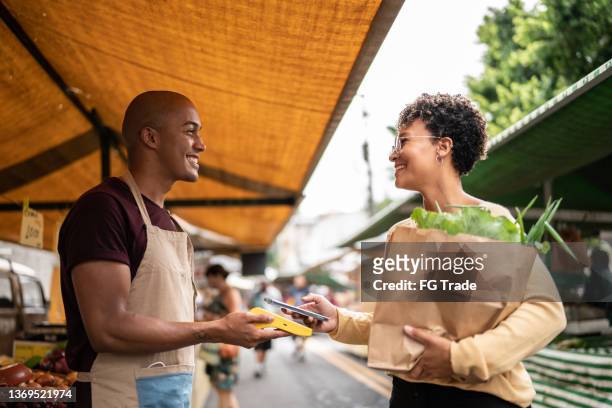young woman paying with mobile phone at a street market - vendor payment stock pictures, royalty-free photos & images