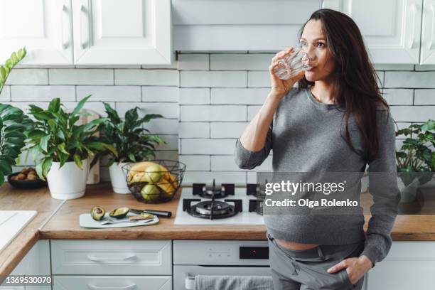 beautiful dark haired pregnant woman wearing grey drink glass of water at kitchen - wasser trinken zu hause stock-fotos und bilder