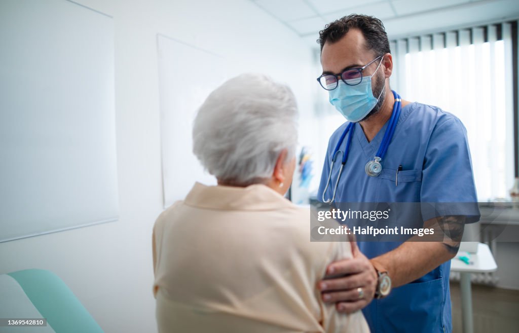 Rear view of senior woman with doctor wearing protective face masks and talking warmly in doctor's office.