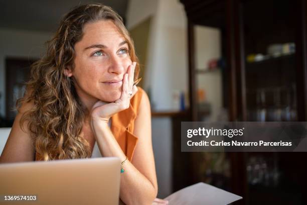 a woman with a dreamy expression working with a laptop from home - dreamers stockfoto's en -beelden