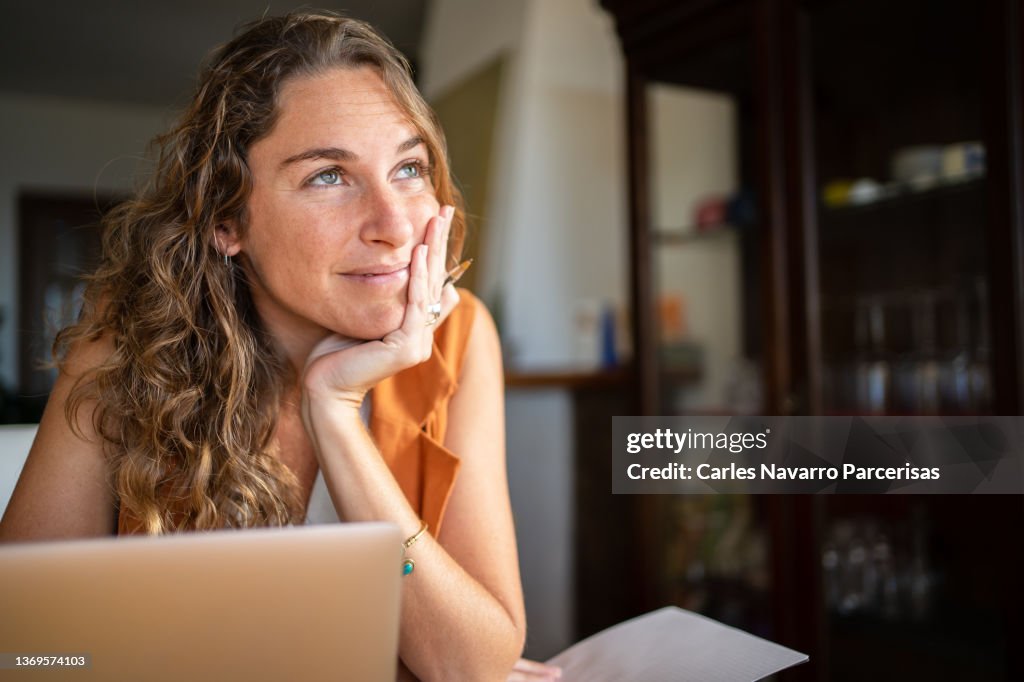 A woman with a dreamy expression working with a laptop from home