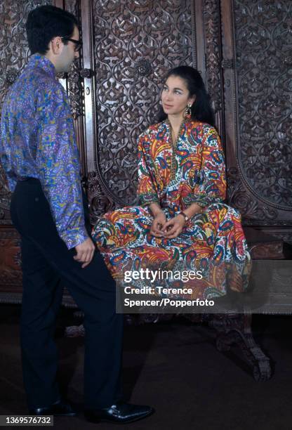 Man and woman in a hippie fashion boutique, the woman wears a kaftan dress in a vibrant floral print, the man wears a purple paisley print shirt,...