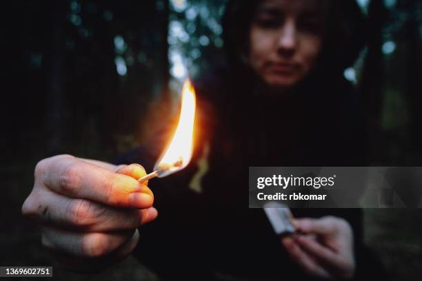 a teenager in a black hood holding a burning match in a dark forest at night. arsonist damages the environment. forest fire danger, flame in the hands of a hooligan - brandstichting stockfoto's en -beelden