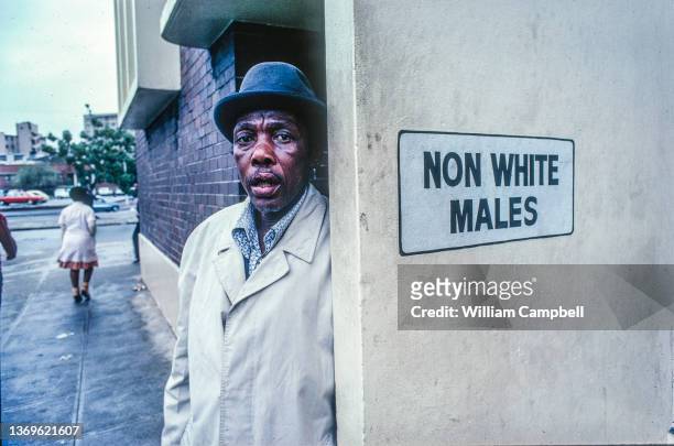 Portrait of an unidentified Blank man, in a hat and overcoat, as he stands beside a restroom sign that reads 'Non White Males,' Johannesburg, South...