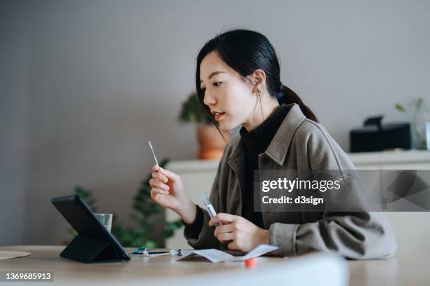 asian woman holding a covid-19 rapid lateral flow test kit, a nasal swab and a plastic tube, carrying out a coronavirus rapid self test at home. she is having a video call appointment on digital tablet with her family doctor for advice and consultation - speed test stock pictures, royalty-free photos & images