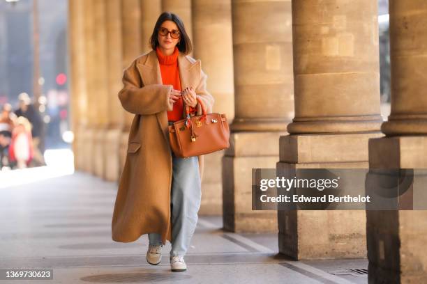 Alexandra Pereira wears brown sunglasses, gold earrings, a neon orange turtleneck wool pullover, a beige oversized long winter coat, a brown shiny...