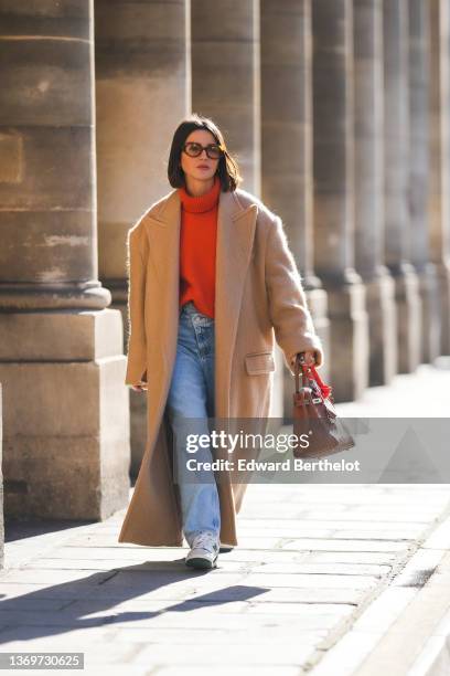 Alexandra Pereira wears brown sunglasses, gold earrings, a neon orange turtleneck wool pullover, a beige oversized long winter coat, a brown shiny...