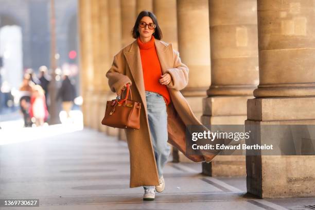 Alexandra Pereira wears brown sunglasses, gold earrings, a neon orange turtleneck wool pullover, a beige oversized long winter coat, a brown shiny...