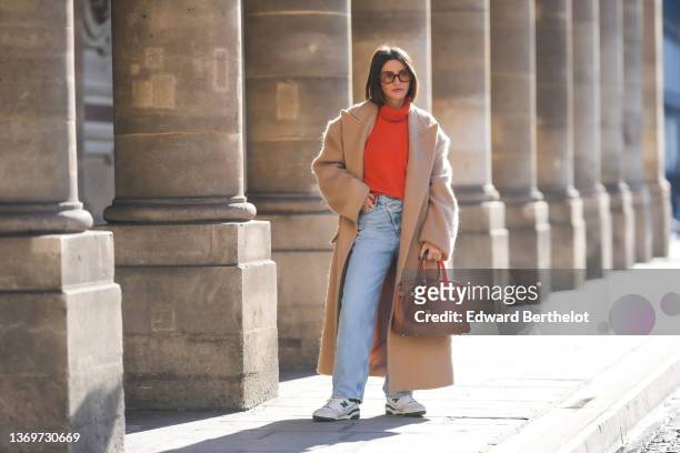 Alexandra Pereira wears brown sunglasses, gold earrings, a neon orange turtleneck wool pullover, a beige oversized long winter coat, a brown shiny...