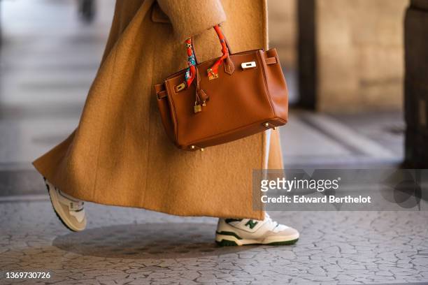 Alexandra Pereira wears a beige oversized long winter coat, a brown shiny leather Birkin handbag from Hermes, an orange / blue / yellow / white print...