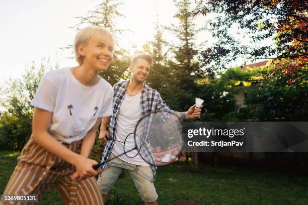 aufgeregtes junges paar, das mit einem freund im vorder- oder hinterhof badminton spielt - playing badminton stock-fotos und bilder