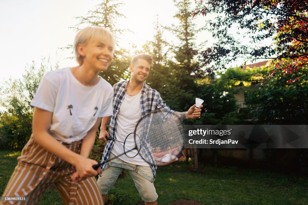 Pareja joven emocionada jugando bádminton con un amigo en el patio delantero o trasero
